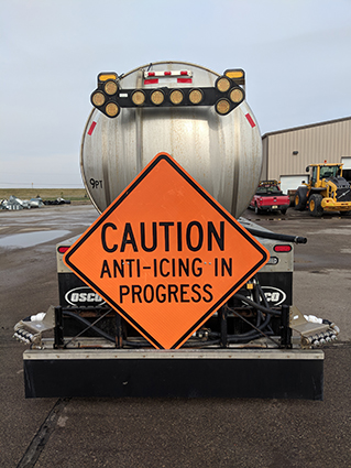 Orange sign on the back of a truck says "Caution Anti-Icing in Process."