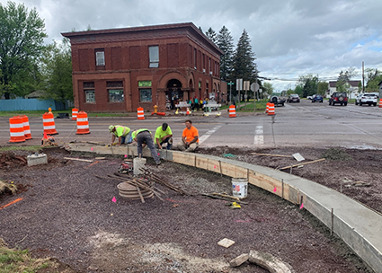 Four construction workers pour concrete for a curb gutter. There is a business behind the work zone.