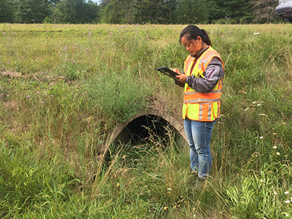 A woman in a safety vest is standing next to a culvert, looking at information on an ipad.