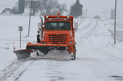 An orange snow plow clears a snow covered road.