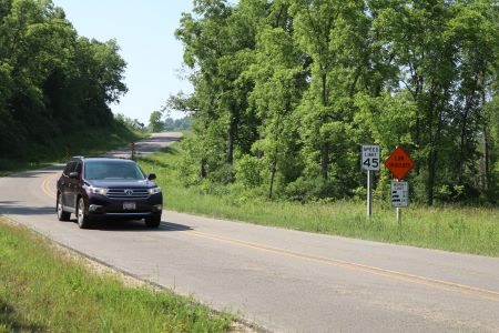 Black SUV driving on a state highway in a rural area. There is a black and white speed limit side on the side of the road. 