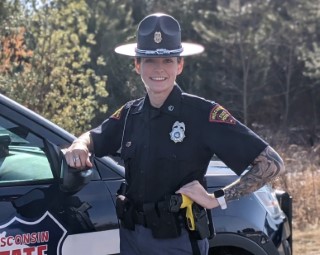 Smiling trooper in uniform leaning on their patrol car.