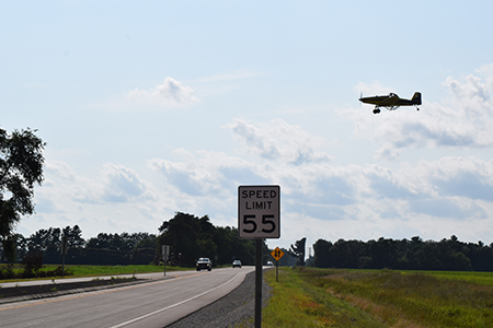 Black SUV driving on a state highway in a rural area. There is a black and white speed limit side on the side of the road.